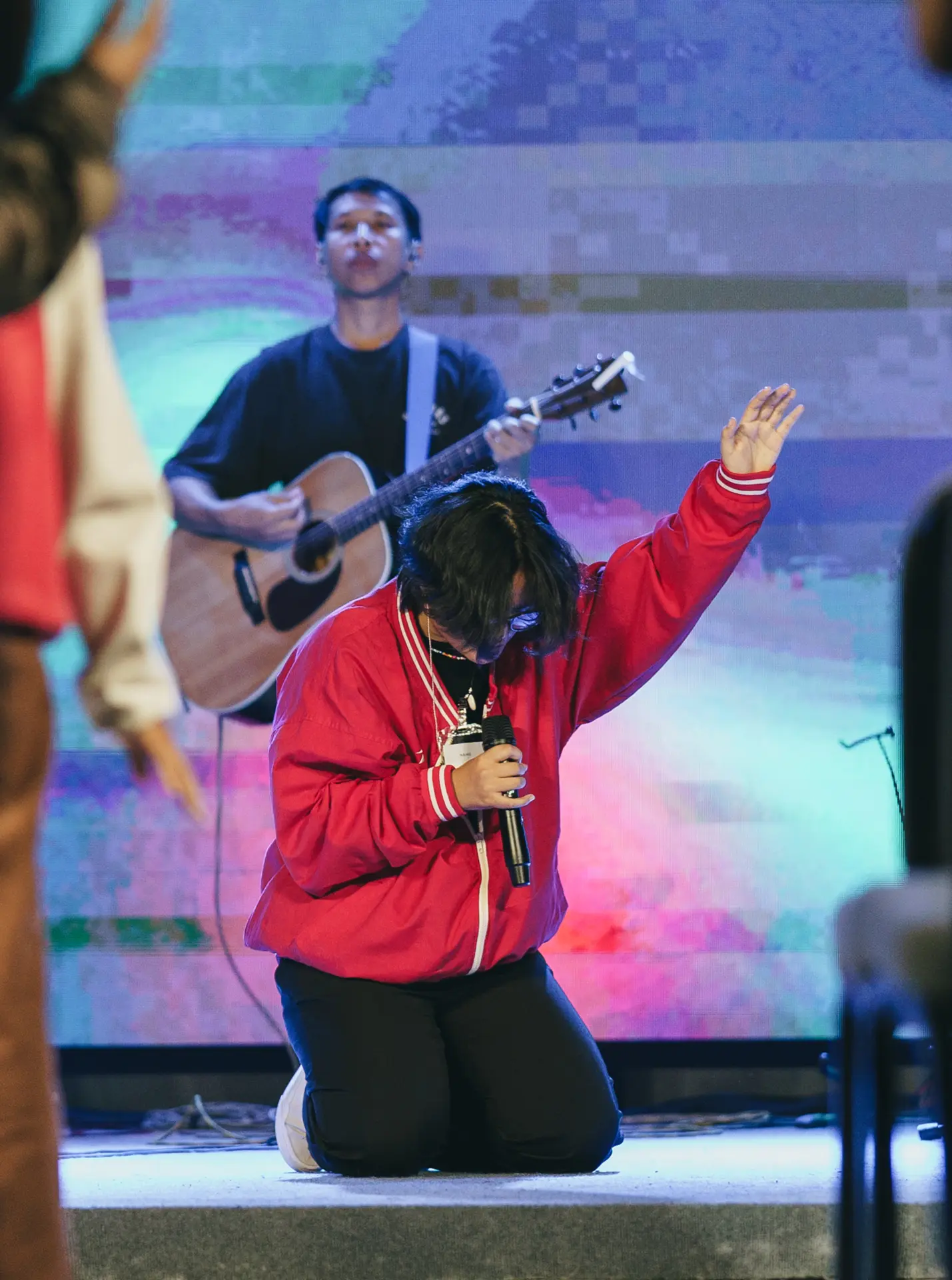 A worshipper in a red jacket bows with one hand raised as a guitarist plays